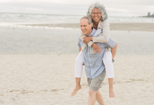Older couple on the beach where one person is giving the other a piggy back as they both smile