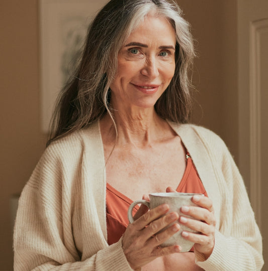 Beautiful, mature woman semi smiling at the camera while holding tea in her hand and wearing the front closing bra for seniors in terracotta