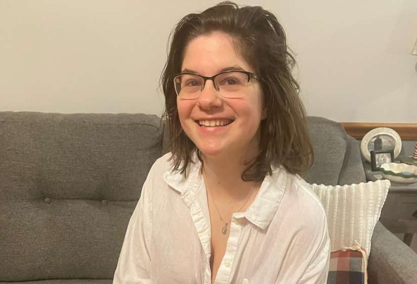 Morgan, a young white woman with a disability smiling widely and happily towards camera while hanging on a couch