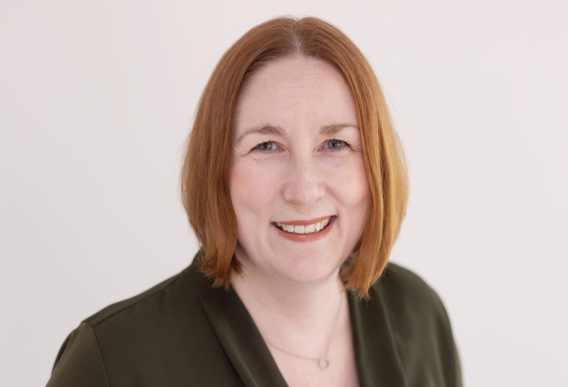 Sarah Dillingham, a white lady with orange-reddish hair smiling at the camera in a dark olive green top