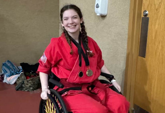 Brie smiling brightly, wearing a red KarateGi while siting in her wheelchair, medals around her neck