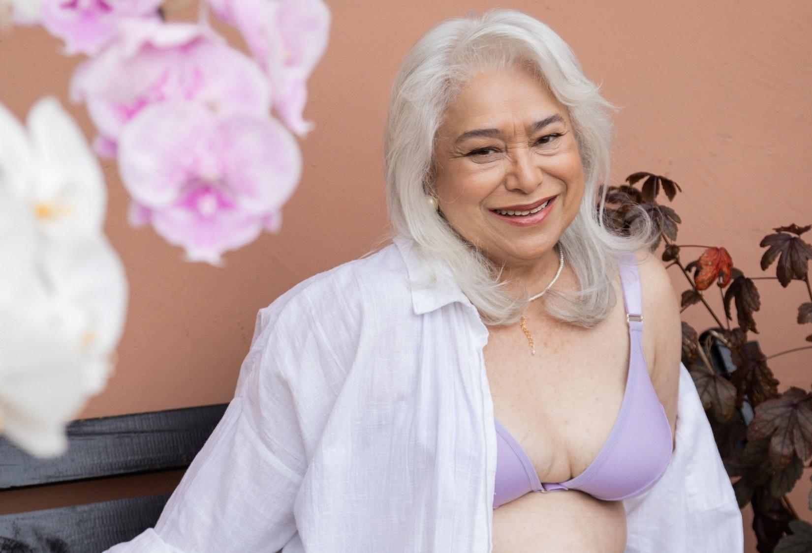 Best Bras for Elderly Ladies: An older woman with long white hair smiles warmly while sitting against a peach-colored wall, wearing the Easy-On Mobility bra with wide straps and a white open shirt.