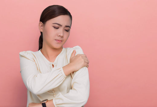 Can’t Fasten Your Bra Because of Arm Pain?: A woman holding her shoulder with a pained expression, indicating arm or shoulder discomfort, against a pastel pink background.