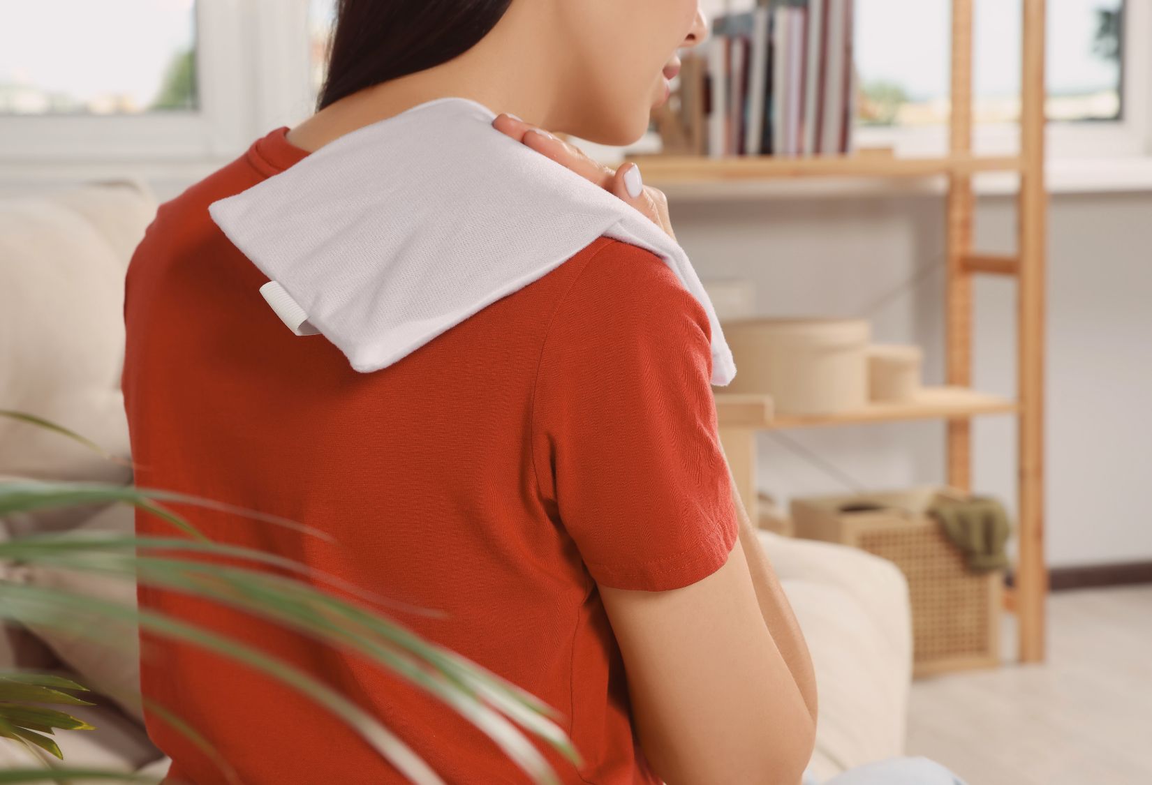Gift Ideas for Someone With a Bad Back: A woman in a red shirt uses a white heating pad on her shoulder while sitting on a couch, suggesting relief from muscle or back pain.