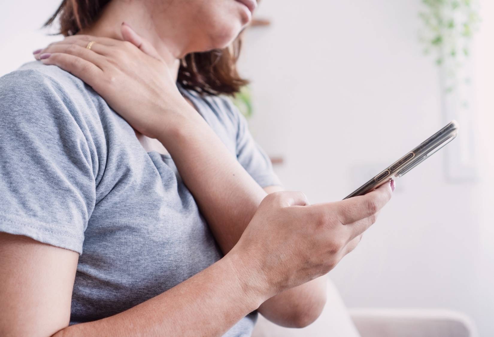 Signs It Might Be Time to Try an Adaptive Bra: Woman rubbing a sore shoulder and neck while looking at her phone