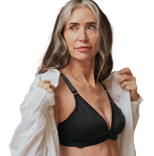 Beautiful, mature woman looking off camera with a confident look while she puts on a white loose button down on top of a black velcro front closure bra for arthritic hands