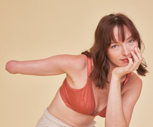 Pretty young woman leaning her face in her hand and raising her other arm parallel to the floor. She has a confident and beautiful expression while staring at camera and showing off her limb difference. She is wearing the one arm bra in terracotta.