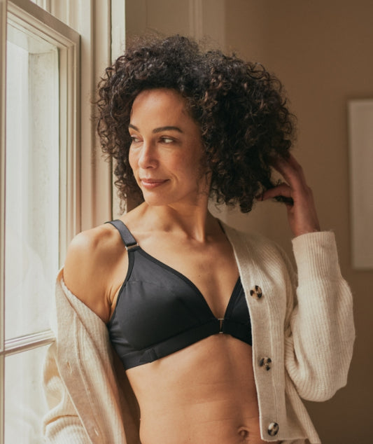 Beautiful, mature woman leaning against a windowsill with a pleased expression while wearing the adaptive wireless front closure bra under a beige cardigan